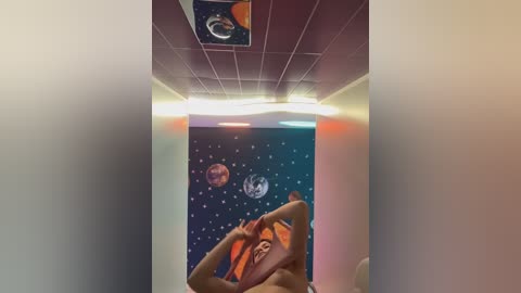 A video of a young woman with fair skin and medium-length, curly brown hair, lying on a bed with her arms above her head, against a starry night sky mural with planets.