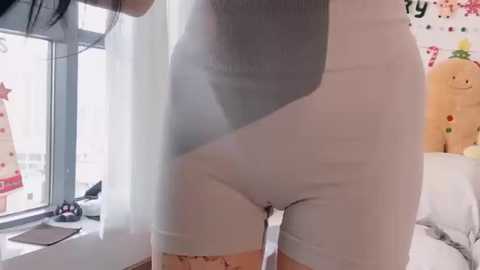 Video of a slender, light-skinned woman wearing tight, white shorts and a grey top, standing in a brightly lit room with a window, plush toys, and decorative items visible.