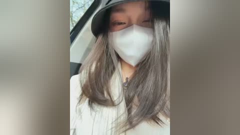 Video of an Asian woman with long, straight black hair, wearing a white face mask, a black wide-brimmed hat, and a beige jacket, partially visible inside a car.