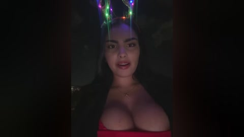 A video of a young woman with large breasts, wearing a red dress, illuminated by colorful, glowing lights on a dark background.