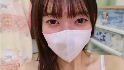 Video of an East Asian woman with straight brown hair, wearing a white surgical mask, in a pharmacy setting. The background shows colorful packaging and medicine shelves.