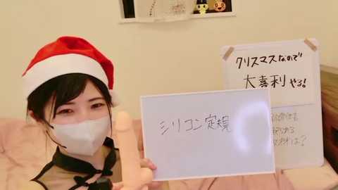 Video of an East Asian woman wearing a red and white Santa hat, a white mask, and a black dress, holding a sign in Japanese in a bedroom with a pink bedspread.