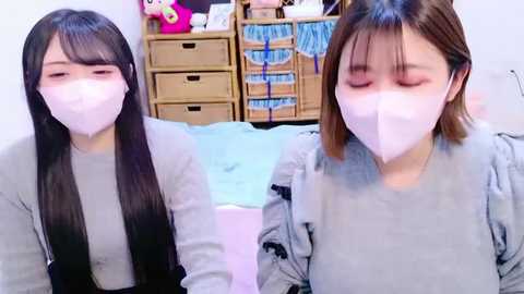 Video of two young Asian women with long black hair, wearing white face masks, gray sweaters, and sitting on a bed in a cluttered room with a wooden cabinet and stuffed toys.