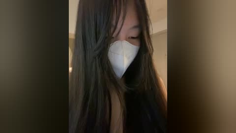 Video of a young Asian woman with long black hair partially covering her face, wearing a white surgical mask and a black top, indoors with blurred beige walls.