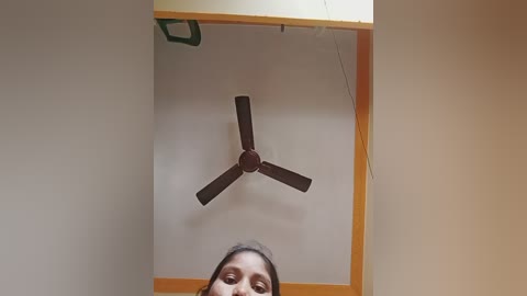 Video of a woman with medium brown skin and long dark hair, standing in a room with a ceiling fan, beige walls, and a wooden door frame.