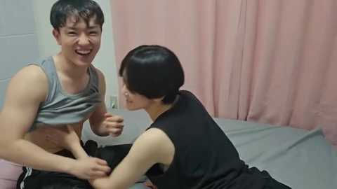 A video of two young Asian men laughing in a medical examination room. The man on the left is shirtless, wearing a gray tank top, while the man on the right, with short black hair, is dressed in black. Pink curtains and a white wall are in the background.