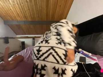 A video of a person in a white and black patterned onesie standing on a bed with a purple sheet, in a room with wooden ceiling and black headboard.