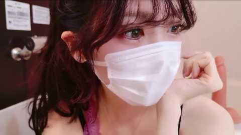 Video of an East Asian woman with long, dark hair, wearing a white face mask and a pink top, holding her hand to her mouth, indoors with a door and papers on the left.