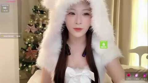 Video of a young Asian woman with fair skin, wearing a white fur hood and white lace top, standing in front of a decorated Christmas tree.