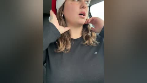 Media: A candid video of a young woman with fair skin, wavy brown hair, wearing a red and white Santa hat and a black sweatshirt, adjusting her hair, seated in a car.