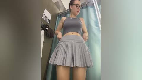 A video of a young woman with fair skin, glasses, and dark hair in a ponytail, wearing a gray sports bra and pleated skirt, adjusting her skirt in a bathroom with teal shower curtain.