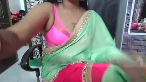 Media: A video of a woman in a bright green sari with a pink bra and matching red leggings, seated on a black office chair, in a room with a brick wall and a mirror.