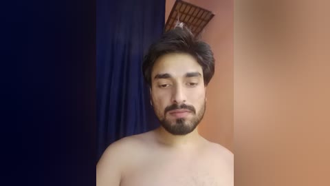 Video of a bearded, light-skinned man with short, dark hair, looking downward, standing against a wall with a dark blue curtain on the left and a peach-colored wall on the right.