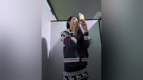 Video of a woman wearing a black and white Nordic-inspired sweater, taking a selfie in a dimly lit, modern bathroom with white walls and a frosted glass door.