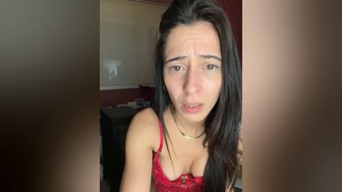 Video of a young woman with long, dark hair, wearing a red lace bra, in a dimly lit room with a red wall and a partially visible black dresser.