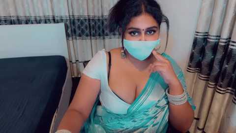 A video of a South Asian woman with medium skin tone and dark hair in a bun, wearing a turquoise sari, blue face mask, and multiple bracelets. She sits on a bed with striped curtains in the background.