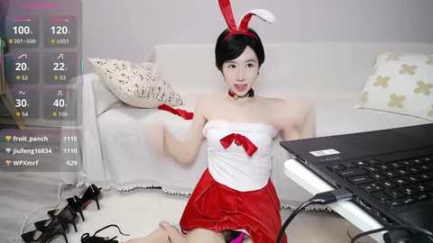 A video of an Asian woman in a red bunny costume, sitting on the floor, using a laptop, with scattered clothes and shoes nearby.