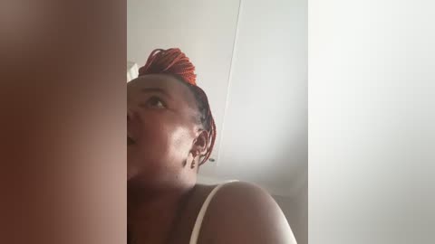 A video shows a Black woman with braided red hair, wearing a tank top, standing in a dimly lit room with a white wall.