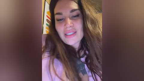 Video of a young woman with long, wavy brown hair, smiling, wearing a light pink hoodie, standing indoors with blurred background featuring colorful, abstract art.