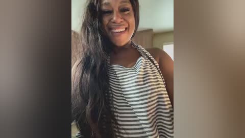 Video of a happy Black woman with long, straight dark hair, wearing a sleeveless, striped top, standing indoors with blurred background.