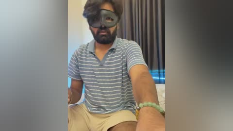 Video of a bearded South Asian man in a VR headset, wearing a striped polo shirt and beige shorts, sitting on a bed in a dimly lit room with dark curtains.