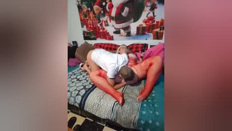 Video of a woman in a red dress lying on a bed with a man in a white shirt and red shorts, surrounded by Christmas-themed decorations and pillows.