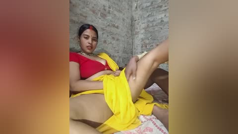 Video of a South Asian woman with medium skin tone and a slender build, wearing a red top and yellow sari, seated on a bed with a red background, displaying an intimate moment with a man.