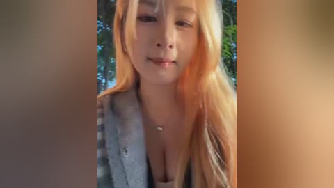Media: Video of a young, light-skinned woman with long, straight blonde hair, wearing a low-cut, patterned top revealing her ample cleavage. She smiles slightly, with a blurred, sunlit outdoor background.