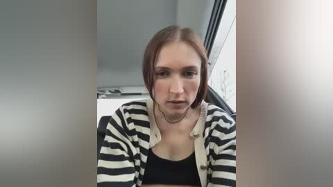 Media: Video of a young woman with light skin and straight, shoulder-length brown hair, wearing a black tank top and a striped cardigan. She looks serious, seated indoors with a window and grey walls behind her.