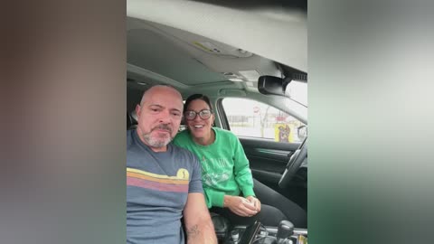 Video of an elderly couple, a bald man with a mustache in a blue shirt, and a woman with glasses in a green shirt, smiling in a car.