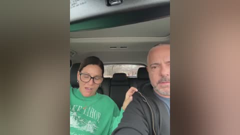 Video of a woman with glasses in a green hoodie and a man in a black jacket seated in a car, with a blurred background.