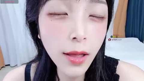 A close-up video of an Asian woman with smooth, fair skin, long black hair, and closed eyes, wearing a black lace top. She has a subtle, natural makeup look. The background shows a bed with white and blue sheets.