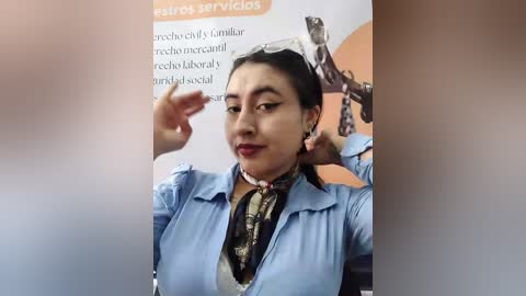 Video of a woman with medium skin tone, dark hair, wearing a light blue shirt, red lipstick, and a patterned scarf, adjusting sunglasses in a blurred background with text.