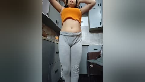 Video of a woman with light skin and medium build, wearing a tight orange crop top and grey sweatpants, standing in a modern kitchen with grey cabinets and a marble countertop.
