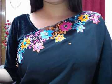 Video of a woman with long black hair wearing a black velvet sari adorned with colorful embroidered floral patterns in pink, blue, yellow, and green. Background features red curtains and a wooden frame.