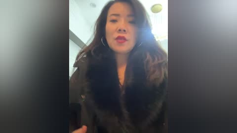Video of an East Asian woman with long dark hair, wearing a black fur coat, red lipstick, and large hoop earrings, looking slightly off-camera in a dimly lit room.