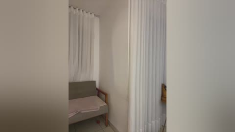 A video of a small, sparsely furnished room with white walls, a bed with a gray cushion and pink blanket, and a white curtain covering a window. A wooden door is partially open to the right.
