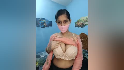 Video of a South Asian woman in a room with blue walls, wearing a pink mask, light pink bra, and a pink shirt, lifting shirt to reveal large breasts, floral wall art, and a bed with patterned bedding.