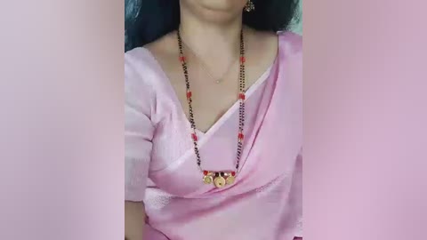 Video of a woman with light skin, wearing a pink saree with a black blouse, adorned with a red-beaded necklace and golden pendant, her blue hair visible in the background.