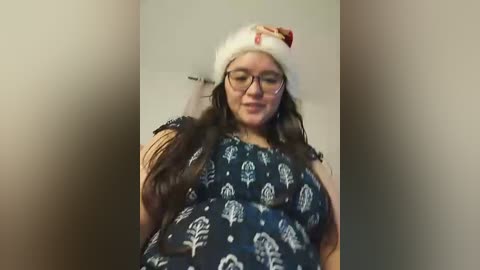 A candid video of a plus-sized woman with long brown hair, wearing a white Santa hat, glasses, and a black top adorned with snowflakes. She has a hand on her head, appearing distressed. The background is blurred, likely indoors.