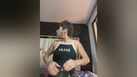 Video of a woman with short, dark hair, wearing a black \"Mama\" tank top, standing in a bathroom with a tiled wall and a towel hanging on the door.