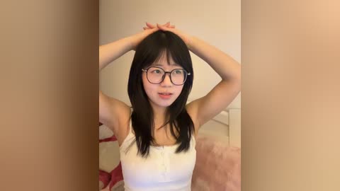 A video of an East Asian woman with long black hair and glasses, wearing a white tank top, standing with arms raised, against a beige wall in a bedroom.