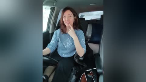 Video of a smiling Asian woman with shoulder-length brown hair, wearing a light blue shirt and black pants, seated in a car with dark interior.