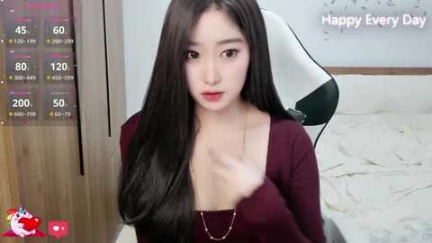 Media: A video of an East Asian woman with long, straight black hair and fair skin, wearing a low-cut burgundy top, sitting on a white gaming chair. Background includes a wooden cabinet and a \"Happy Every Day\" message.