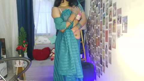 Media: Video of a woman in a teal off-shoulder saree, accessorized with bangles, standing in a colorful, cozy room with a red heart pillow, framed photos, and a glass table.