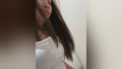 A close-up video of a woman with long, straight brown hair, wearing a white tank top, captured from a low angle, with a blurred background.