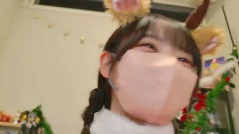 A blurry video of an Asian woman in a cat costume with a white mask, wearing a white dress and braided hair, in a festive room with Christmas decorations.