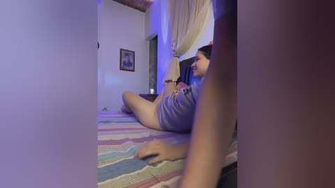 A video of a young woman with medium skin tone, dark hair, and a slender build, lying on a striped bedspread, wearing a grey shirt, in a dimly lit bedroom with a framed picture on the wall.