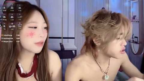 Video of two Asian women with pale skin, one with long brown hair and the other with short blonde hair, both topless and wearing makeup. Background shows a modern indoor setting.