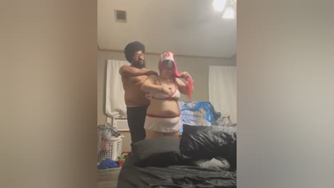 Video of a shirtless man with a beard, wearing dark pants, embracing a busty woman in a nurse costume, standing in a cluttered bedroom.